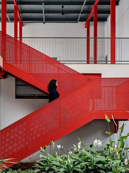 A person pauses on the landing of the vibrant red staircase, which acts as the central spine of the building. The open-tread design and perforated panels create a feeling of lightness and transparency.