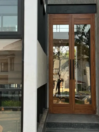 The entrance of the 6AM House, featuring simple, elegant wooden doors that contrast with the raw, modern structure. The reflection in the glass captures the surrounding environment, integrating it into the facade.