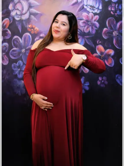 A happy portrait of a mother-to-be in a simple red dress, pointing to her baby bump against a vibrant floral backdrop in our studio.