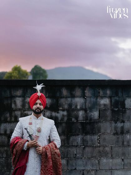 A powerful portrait of the Sikh groom in his traditional attire, holding a kirpan. The dramatic sky and strong pose create a majestic and timeless image.