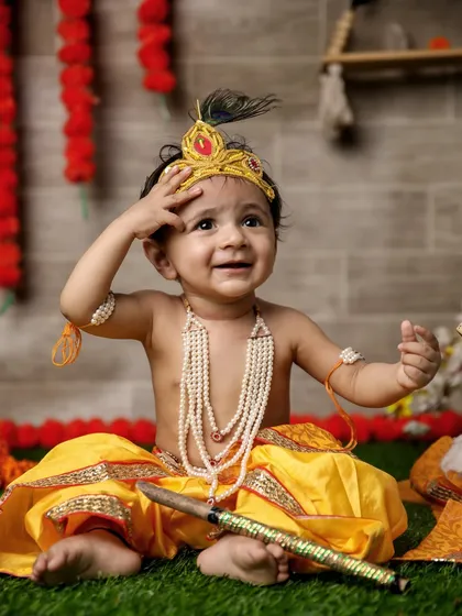 A playful, candid shot of a baby Krishna looking up with a curious and happy expression. The artificial grass and flower garlands add to the festive, outdoor feel of this indoor setup.