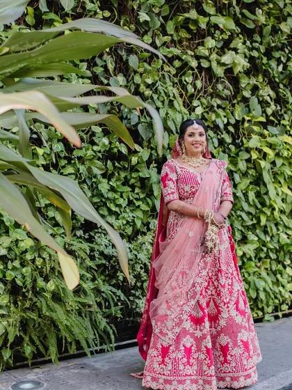A classic red lehenga look for a destination wedding. The makeup is full bridal glam, with a dewy finish and a soft sculpt to complement the rich tones of her outfit against a beautiful green backdrop.