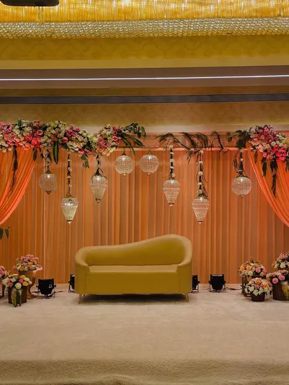 This elegant stage setup doubles as a beautiful photo opportunity. The soft peach drapes, floral arrangements, and hanging glass lamps create a romantic and sophisticated backdrop.
