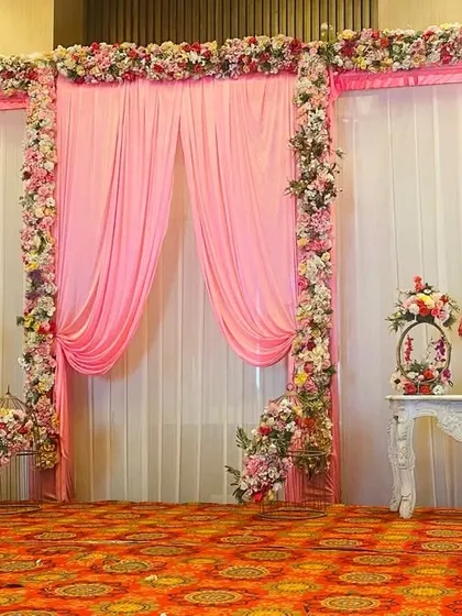 A classic and elegant stage setup perfect for a golden jubilee anniversary celebration. The design uses soft pink and white drapes, framed by floral arrangements, with vintage-style tables for a timeless feel.
