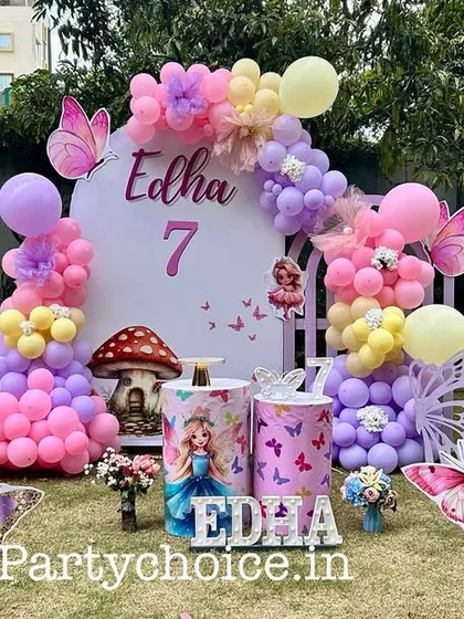 An outdoor butterfly-themed birthday setup for a 7th birthday. The decor uses a pastel balloon arch, butterfly cutouts, and themed plinths, creating a lovely garden party atmosphere.