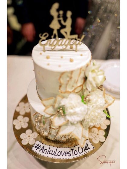 A close-up of an elegant two-tiered engagement cake, featuring delicate wafer paper flowers and a custom hashtag, seen here at the celebration.