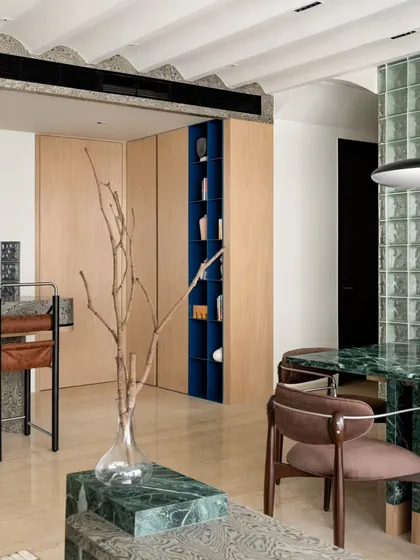 A view from the dining area towards the living room, showing the cohesive use of materials like green marble and light wood veneer. The open-plan design encourages connection and flow between spaces.