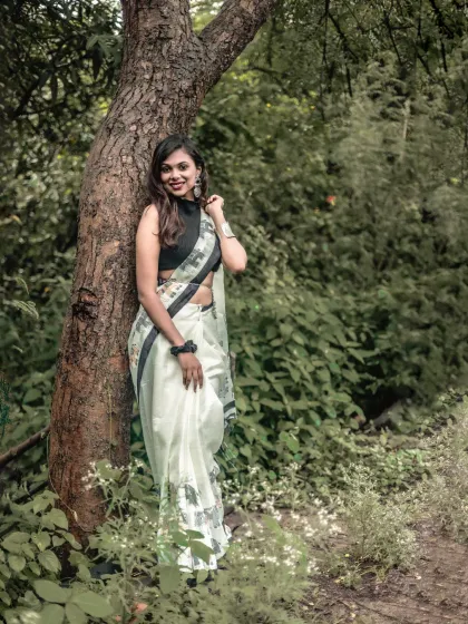 A full-length portrait of a woman in a stylish saree, leaning against a tree. The composition balances the subject with the natural environment.