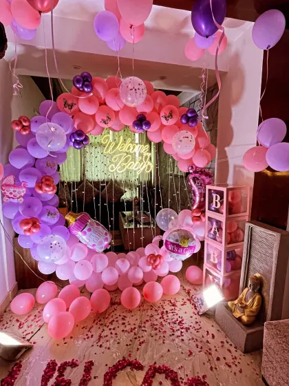 A grand 'Welcome Baby' decoration for a home entrance. This setup includes a crescent-shaped balloon arch in pink, purple, and lavender, a 'Welcome Baby' neon sign, and the floor decorated with rose petals spelling 'WELCOME'.