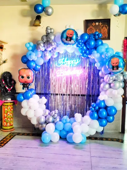 A Boss Baby themed birthday decoration in a home setting. The circular backdrop is framed with blue, white, and silver balloons and features a shiny fringe curtain and a neon sign.