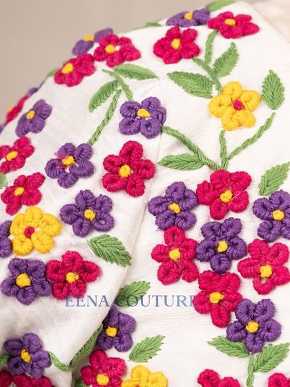 A garden of flowers on a blouse. This white raw silk piece is covered in vibrant, multi-colored floral French knot embroidery, making it a cheerful and eye-catching addition to any festive wardrobe.