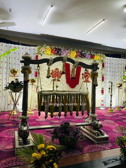 A traditional naming ceremony setup with a decorated cradle placed before a backdrop of white flowers. The central 'Namam' symbol adds a spiritual element to the decor.