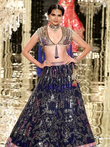 A full-length view of a model on the Suneet Varma runway. The makeup was designed to complement the intricate, multi-colored embroidery of the lehenga, with a focus on a flawless base and defined eyes.