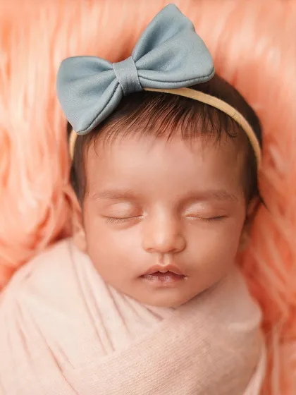 A close-up of a newborn girl with a blue bow, sleeping on a soft, peach-colored fur. The color combination is gentle and sweet, perfect for a baby girl's portrait.