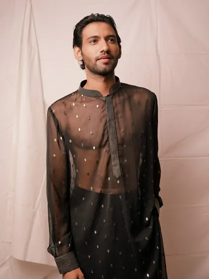 A standing portrait of a male model in a modern, sheer black kurta with silver polka dots. The simple white background and clean lighting make this a perfect shot for an e-commerce product listing.