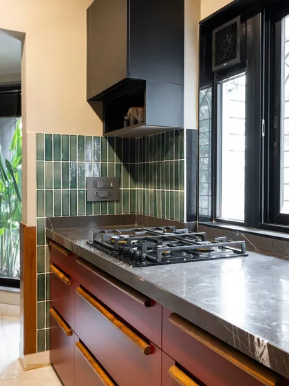 A compact yet characterful kitchen corner. The deep red cabinets, dark marble, and vertical green tiles create a rich, moody palette that is both sophisticated and unique.