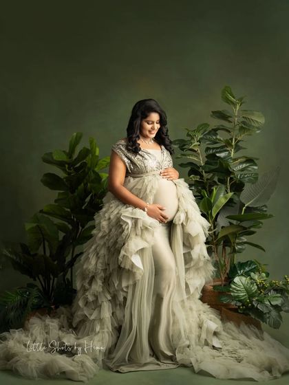 A striking portrait of a mother-to-be in a glamorous, silvery-green ruffled gown. She is surrounded by lush green plants, creating a beautiful, nature-inspired theme.