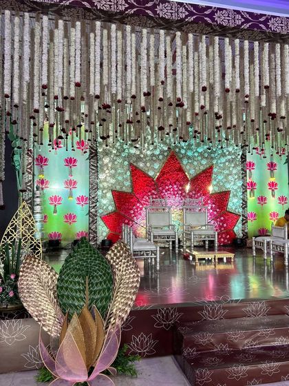 A vibrant and artistic stage with a lotus theme. The backdrop features a large, stylized red lotus and hanging floral strings, creating a colorful and divine setting for a traditional ceremony.