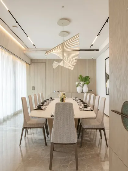 A view from the doorway into the serene dining room, where a long table is set for twelve. The space is defined by its clean lines, neutral palette, and a stunning sculptural light fixture that appears to float above the table.
