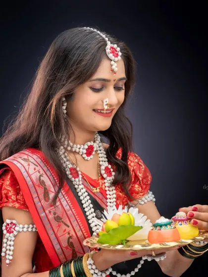 A graceful portrait of a woman in a traditional saree and sugar jewelry, holding a platter of festive sweets against a dark background.