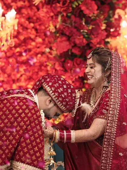 Bowing down for love is the best win. This playful moment during the jaimala ceremony, against a stunning red floral backdrop, is full of joy and adoration.