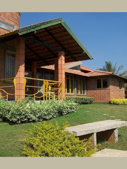 A farmhouse with a welcoming verandah, built with hollow clay blocks and surrounded by a green lawn, embodying relaxed, sustainable living.