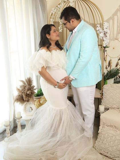A full-length shot showcasing the beautiful white mermaid-style gown. The couple holds hands, sharing a loving gaze in a carefully styled boho studio setting with pampas grass and macrame details.