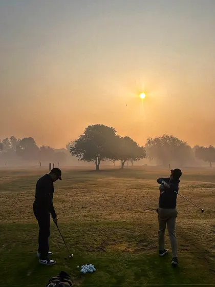 Two golfers practice on the grass tees as the sun rises through the morning mist.