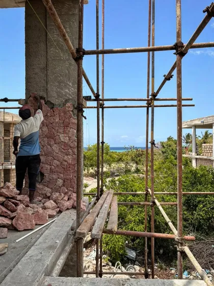 A craftsman stands on scaffolding to apply stone cladding to a column. The view from this vantage point shows the proximity of the site to the ocean, a constant source of inspiration for the design.