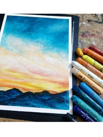 An oil pastel landscape showing a beautiful sunset over mountains, with a box of pastels alongside to show the tools of the trade.