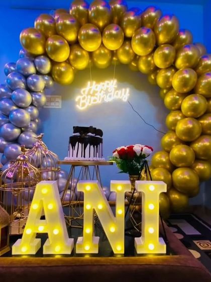A cozy home setup with a gold and silver balloon arch. The display is built around the cake stand and includes decorative birdcages and light-up letters for the name 'ANI'.