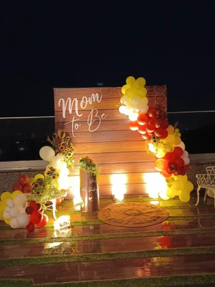 A rustic 'Mom to Be' backdrop with a wooden panel, decorated with a warm-toned balloon garland for a cozy, outdoor baby shower.