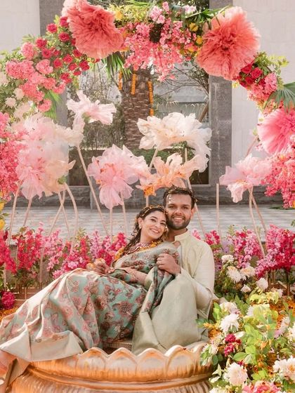 The couple shares a happy moment in their custom Haldi seating, surrounded by a garden of fresh flowers and whimsical decor.