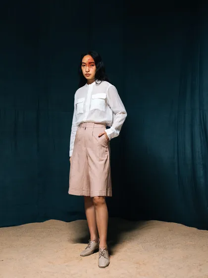 A full-length shot from the same campaign, showing the complete look of a sheer white shirt and tailored shorts. The set includes sand on the floor, adding a textural, earthy element.
