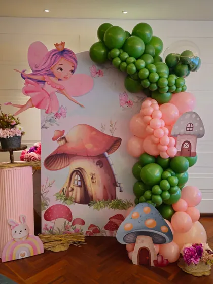 Welcome to a magical fairyland. This enchanting setup for a little one's birthday includes a fairy and mushroom house backdrop, surrounded by a whimsical pink and green balloon garland.