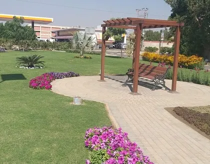 A different view of the public park, showing the seating area with a pergola, surrounded by colorful flower beds and a healthy lawn. This is a space designed for community relaxation and enjoyment.