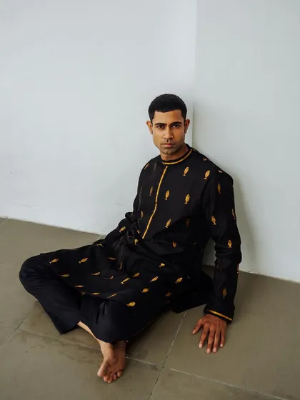 A minimalist shot for the Wabi Sabi menswear lookbook. The model is seated against a plain wall, drawing full attention to the black embroidered kurta.