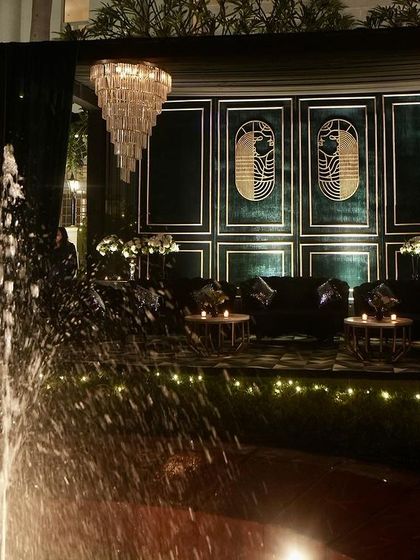 A beautiful view of the Sangeet lounge area, framed by a fountain, showcasing the dramatic chandeliers and sophisticated dark teal decor.