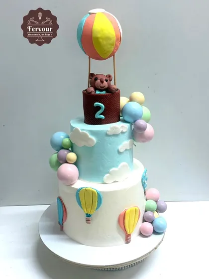 A two-tier hot air balloon theme cake with a teddy bear passenger. This truffle cake is decorated with pastel-coloured balloons and clouds.
