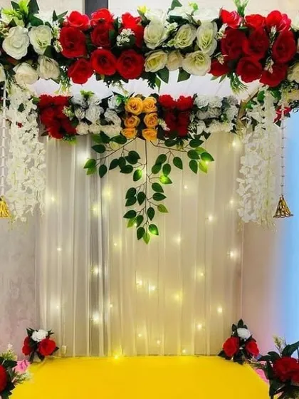 This backdrop features a beautiful arch of red, white, and yellow roses over a curtain of warm fairy lights. The design includes hanging white floral strings and bells, blending modern style with traditional elements.