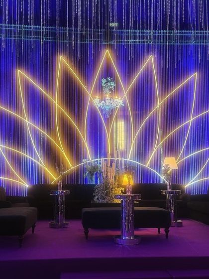A stunning lounge area at the celestial-themed sangeet. A large neon lotus serves as the backdrop, complemented by elegant chandeliers and modern lamps, all under a canopy of deep blue and purple light.