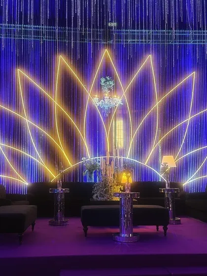 A stunning lounge area at the celestial-themed sangeet. A large neon lotus serves as the backdrop, complemented by elegant chandeliers and modern lamps, all under a canopy of deep blue and purple light.