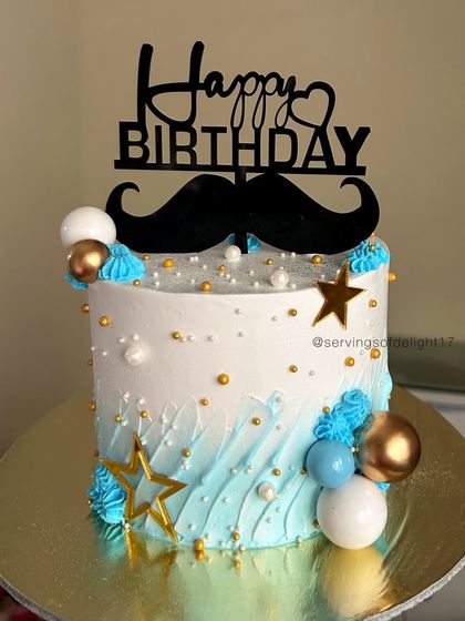 A stylish birthday cake for him, featuring a blue ombre wave design, a mustache topper, and gold and blue chocolate spheres.