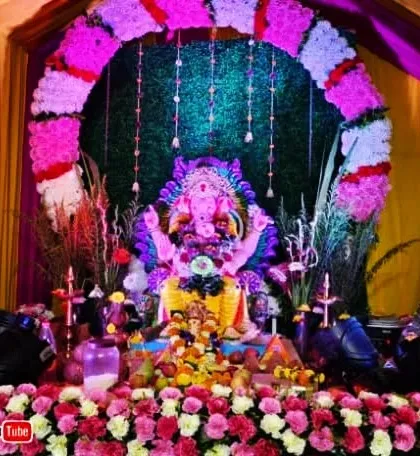 A grand and colorful floral decoration for a community Ganpati pandal. The setup features a large arch made of artificial flowers and a beautifully decorated stage for the idol.