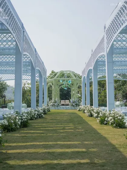 The long aisle leading to the green mandap, flanked by white lattice archways that create a beautiful, structured pathway for the wedding procession.