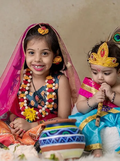 A heartwarming photo of two siblings dressed as Radha and Krishna. The older sister smiles at the camera while her baby brother chews on his flute.