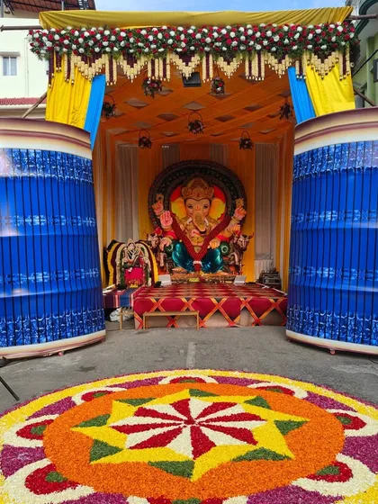 This is a large-scale outdoor pandal we constructed for a Ganesh Chaturthi festival celebration. The setup includes a decorated stage for the idol, vibrant yellow and blue fabric draping, and a massive, colorful flower rangoli on the ground, perfect for community and corporate festive events.