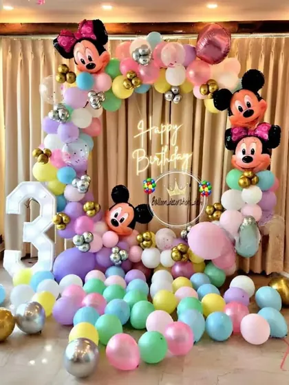 A colorful Mickey and Minnie Mouse theme for a third birthday. The setup includes a pastel balloon arch with character heads, a "Happy Birthday" neon sign, and a large light-up number '3'.