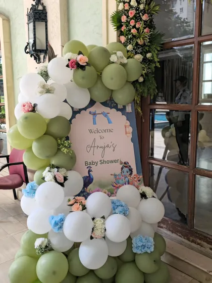 A welcome sign and balloon arrangement for a Krishna-themed baby shower. The entrance features a custom sign for 'Anuja's Baby Shower' with Krishna graphics, framed by an organic balloon garland in sage green and white with floral accents.