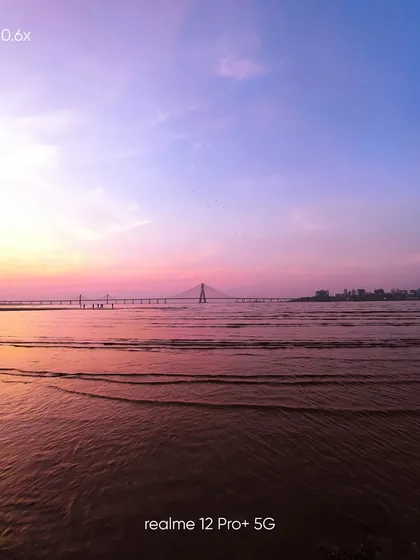 An ultra-wide 0.6x mobile photo that captures the full length of the Bandra-Worli Sea Link at sunset, emphasizing the scale of the engineering marvel against the natural beauty of the coast.